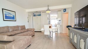 A living room with a beige sofa, a dining area with a white table and chairs, and a kitchen in the background.
