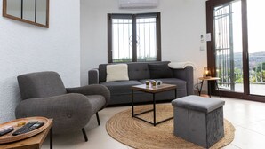 A modern living room with a grey sofa, a grey armchair, a small coffee table, and a grey ottoman. There is a large window with a view of greenery and a balcony. - Wonderful private villa for 6 people with private pool, WIFI, A/C, TV and terrace (Calonge)