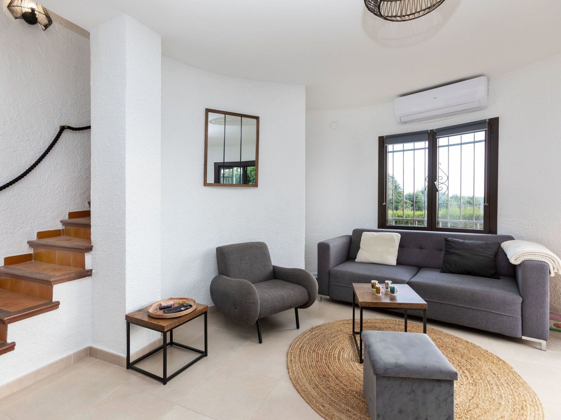 Una sala de estar con un sofá gris, una pequeña mesa de madera, un sillón gris, un tapiz tejido y una escalera con escalones de madera.