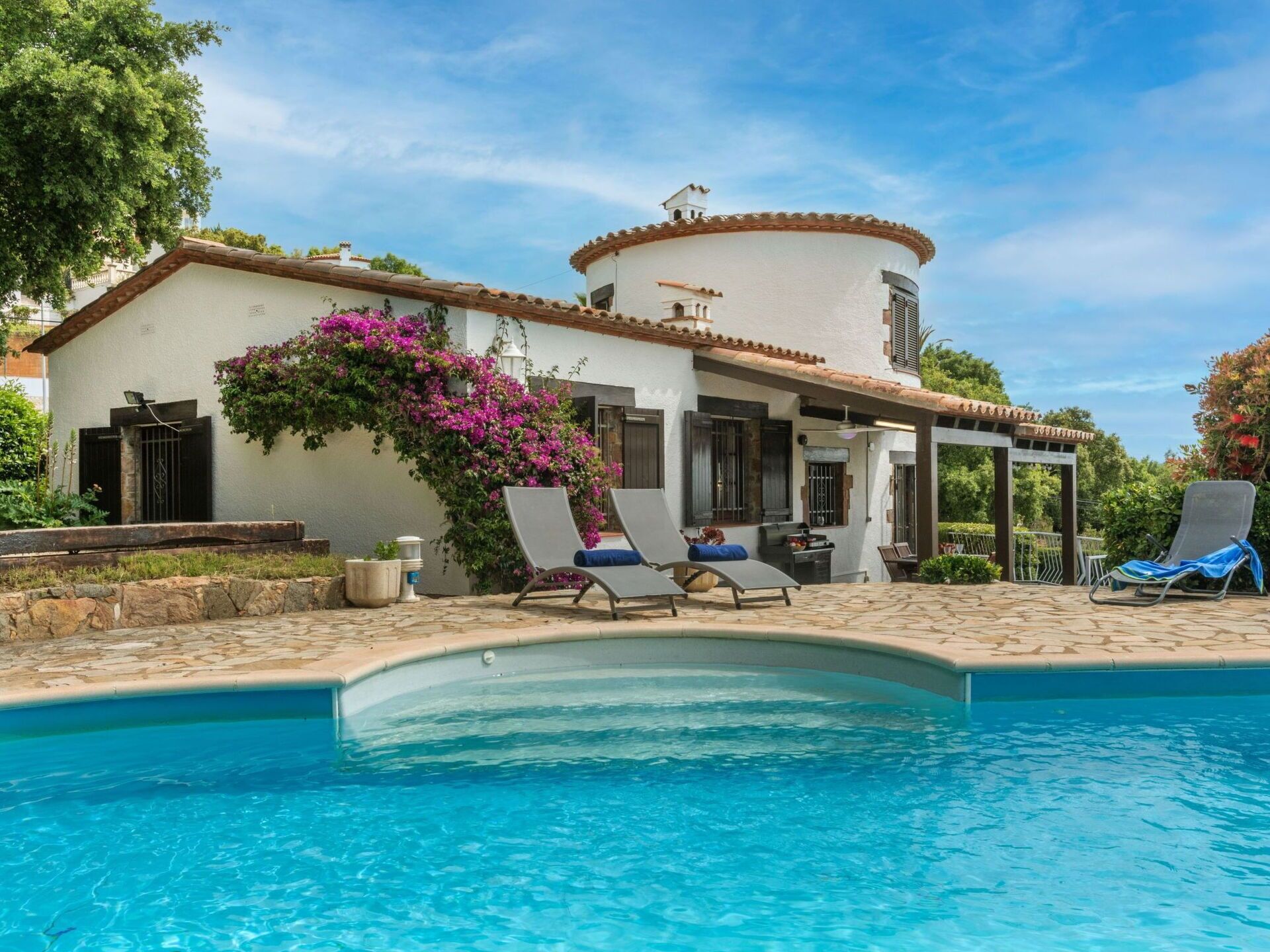 Un espace aménagé au bord de la piscine, avec des chaises longues, une pergola et un bâtiment blanc surmonté d’une tour ronde.