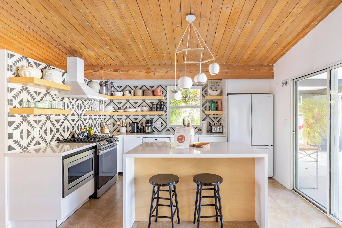 A modern kitchen with a wooden ceiling, white cabinetry, and a patterned backsplash. There are two stools at the island and a hanging light fixture above.