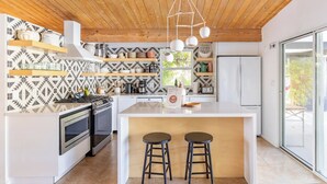 Eine moderne Küche mit Holzboden, weißen Schränken und einer gestreiften Spritzschutzwand. An der Kücheninsel stehen zwei Hocker und darüber hängt eine Leuchte.