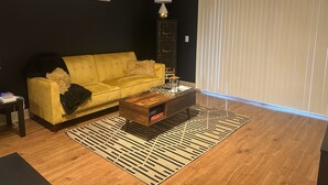 A living room with a yellow sofa, a wooden coffee table, a patterned rug, a lamp, and a framed artwork on the wall. - Cool haven in the city, close to the medical center and NRG. (Houston)