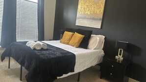 A bedroom with a bed, a nightstand, a lamp, a painting, and a window with blinds. - Cool haven in the city, close to the medical center and NRG. (Houston)