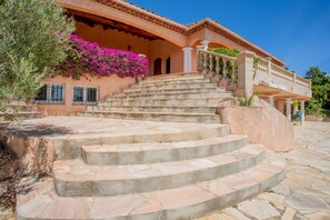 A building with a stone staircase leading to a balcony, surrounded by greenery and a clear sky.