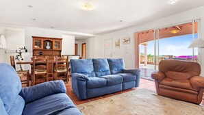 Een woonkamer met een blauwe bank, een bruine fauteuil, een houten eettafel met stoelen, en een groot raam met uitzicht.