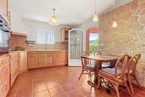 A kitchen with wooden cabinets, a dining table with chairs, a stone wall, and a window.