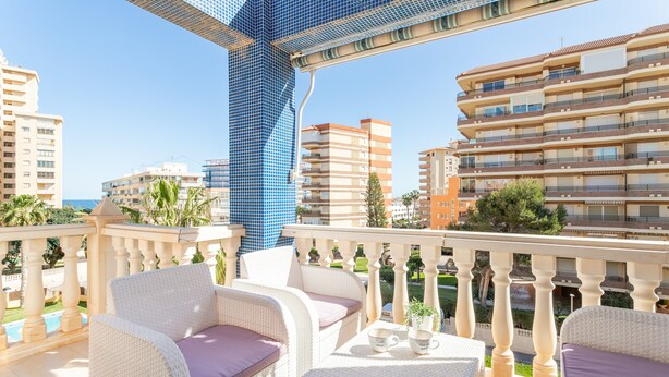 Un balcón con mobiliario de mimbre, una piscina y vistas de edificios circundantes.
