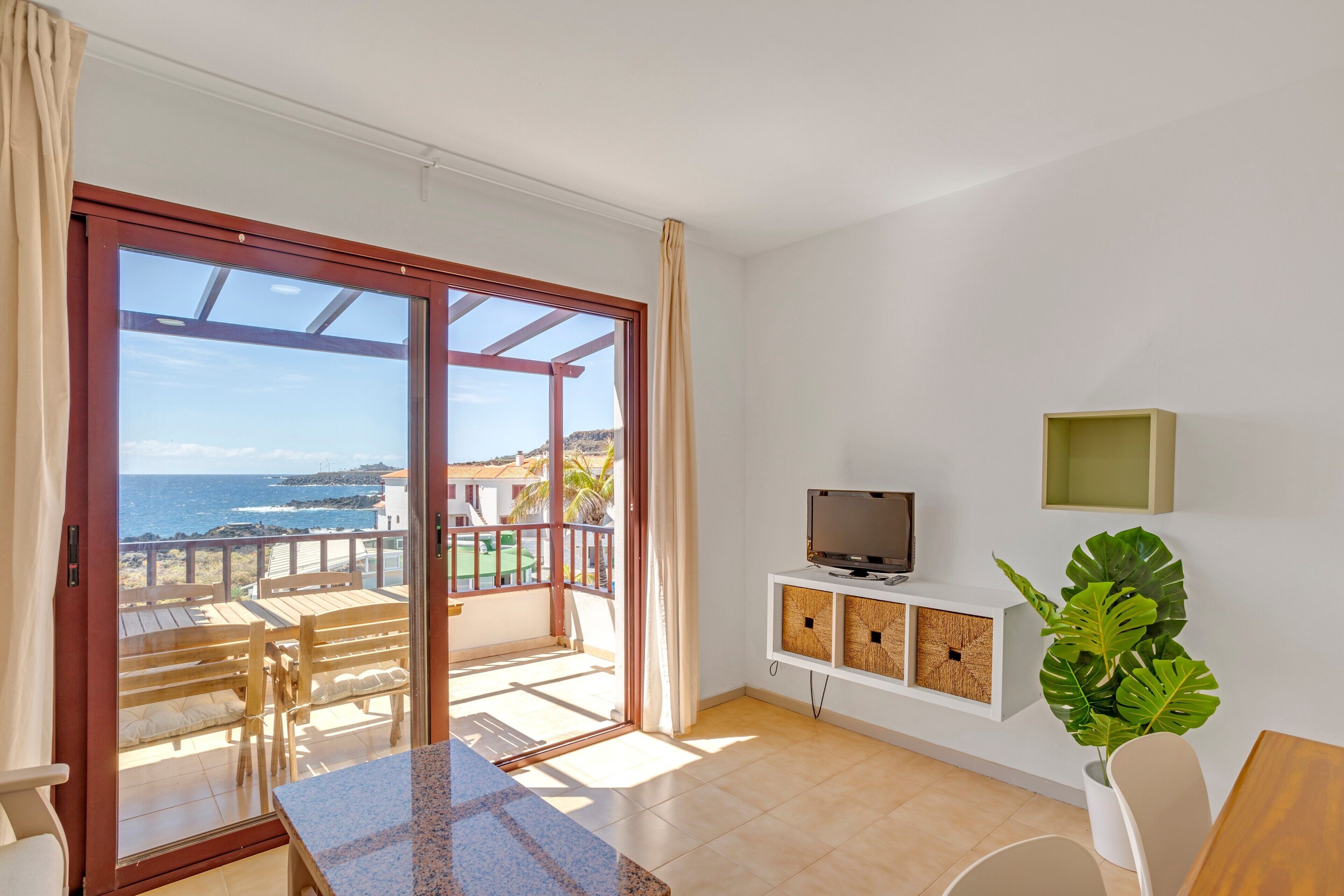 Ein Zimmer mit einer Schiebetür zu einem Balkon mit Meerblick. Ein Fernsehgerät steht auf einem weißen Schrank, daneben ein Blumentopf und ein Holztisch.