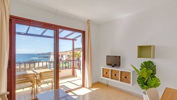 Ein Zimmer mit einer Schiebetür zu einem Balkon mit Meerblick. Ein Fernsehgerät steht auf einem weißen Schrank, daneben ein Blumentopf und ein Holztisch.
