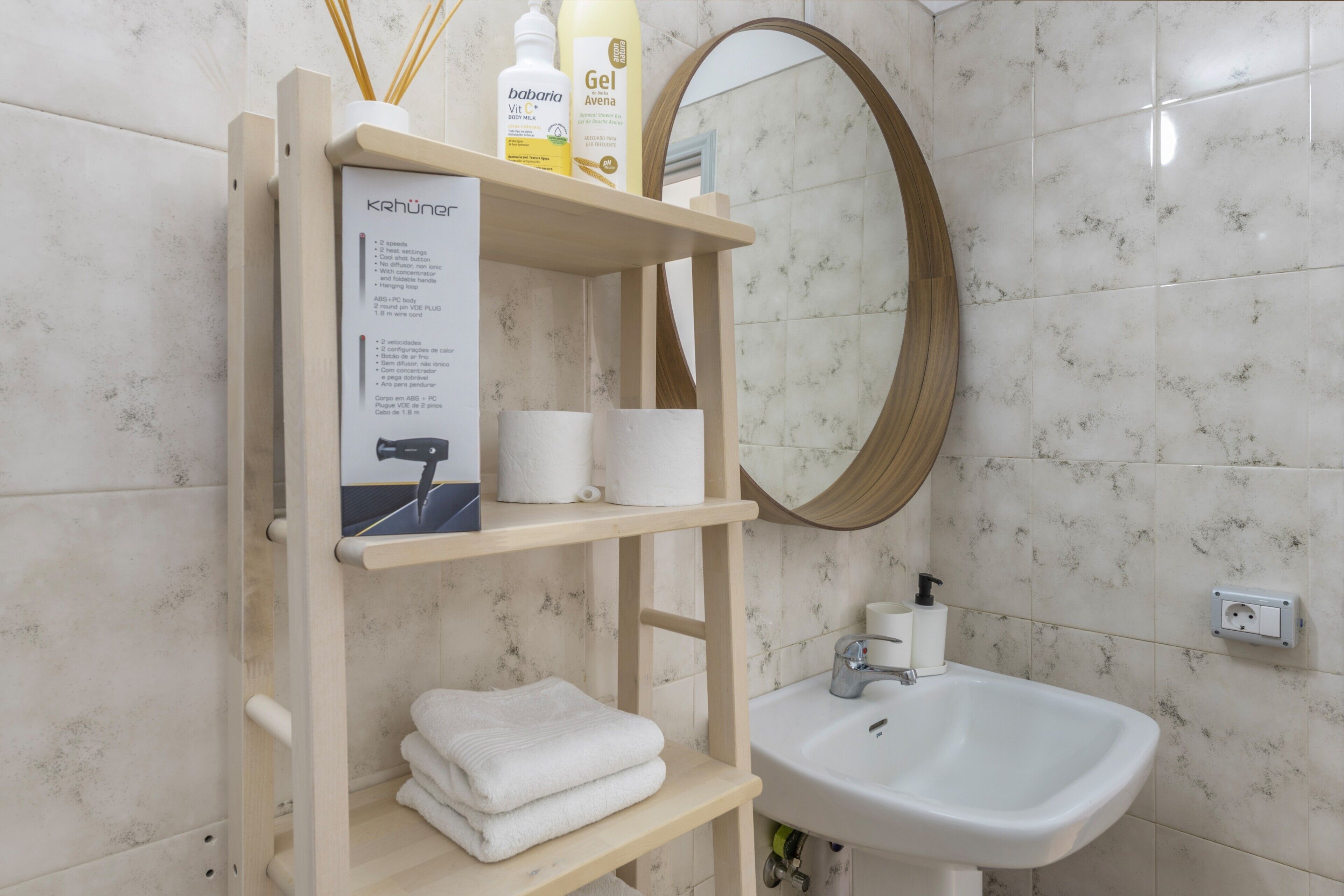 Une salle de bain avec un mur recouvert de carreaux de marbre, une étagère en bois où sont posés des articles de toilette, un lavabo blanc et un miroir.