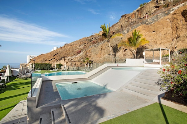 A pool area with multiple swimming pools, a grassy area, and a rocky hillside in the background.