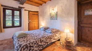 A bedroom with a wooden ceiling, a bed with patterned bedding, a nightstand with a lamp, and a door with a wooden panel. - La Toumet – Family Home with Pool and Pyrenees View (MALABAT)