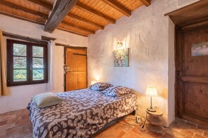 A bedroom with a wooden ceiling, a bed with patterned bedding, a nightstand with a lamp, and a door with a wooden panel. - La Toumet – Family Home with Pool and Pyrenees View (MALABAT)