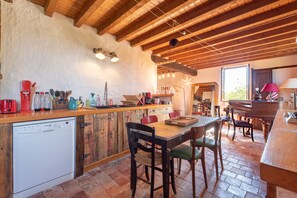 A rustic kitchen with wooden beams, a dishwasher, a dining table with chairs, and a piano in the background. - La Toumet – Family Home with Pool and Pyrenees View (MALABAT)