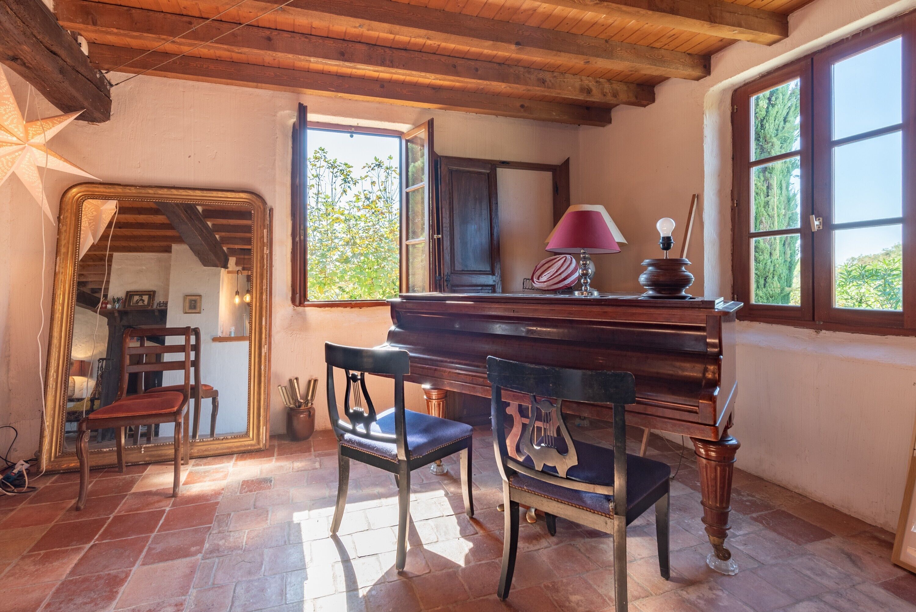 A room with a piano, two chairs, a mirror, and a window with a view of trees.