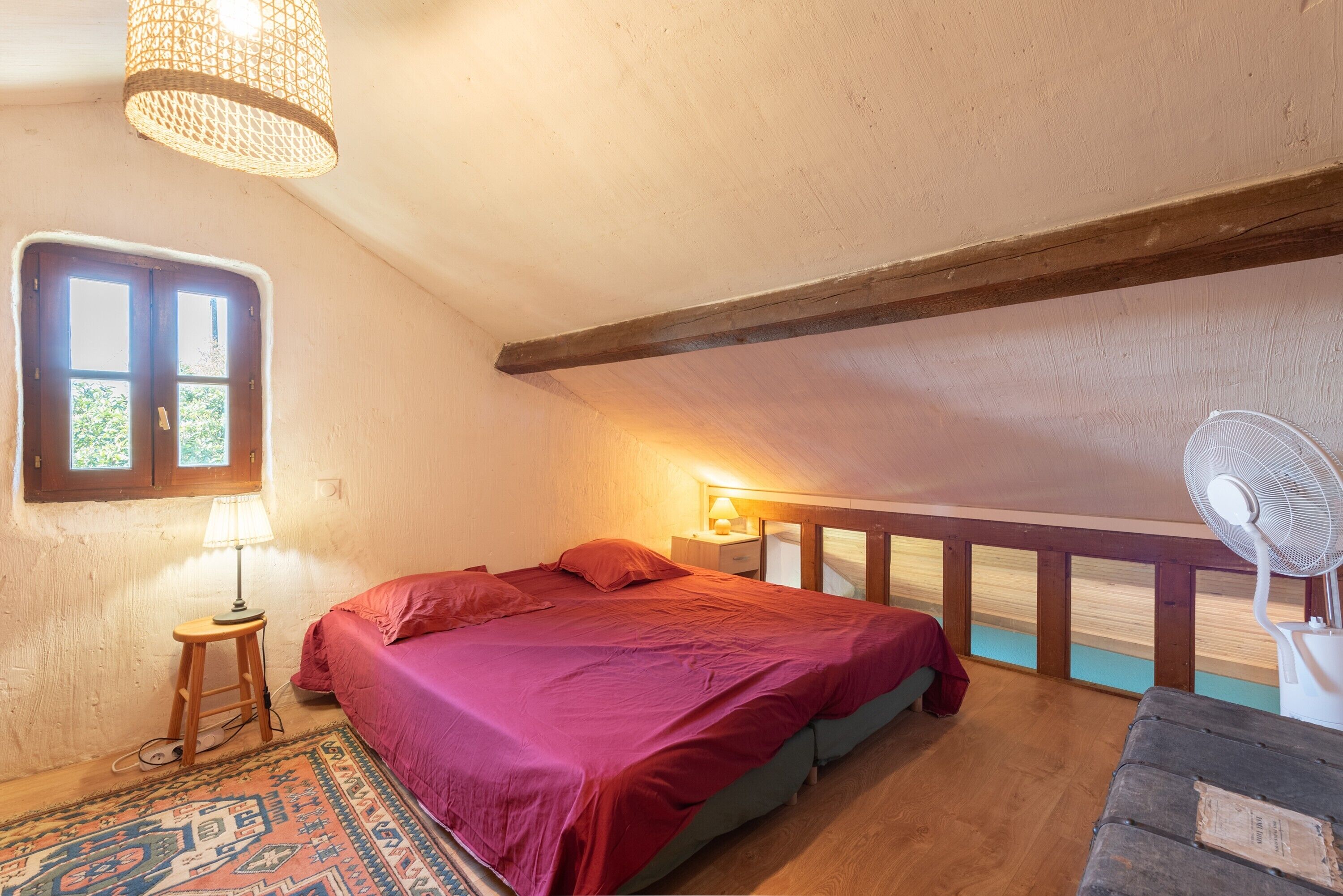Quarto com cama, luminária, ventilador, janela e viga de madeira no teto.