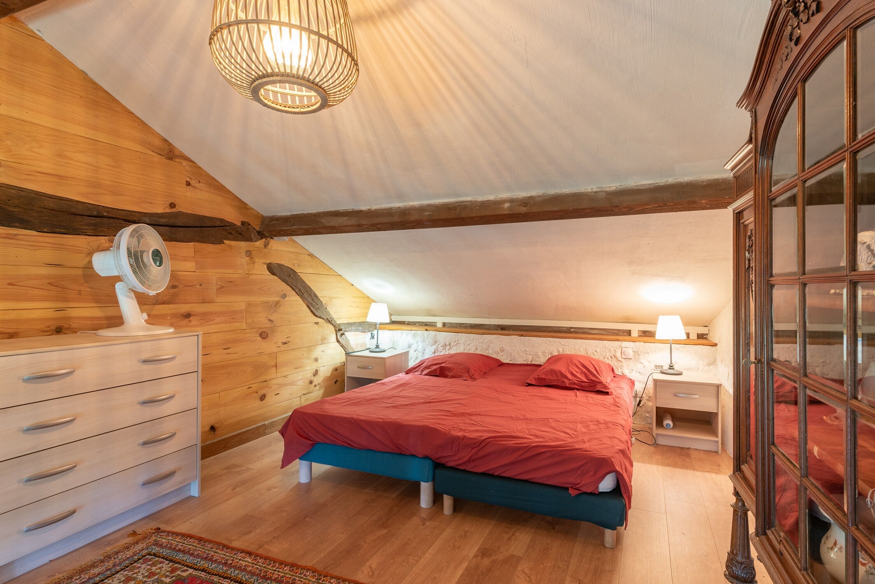 Une chambre avec un plafond en bois, un lit avec une literie rouge, une table de chevet avec une lampe et une commode avec des tiroirs.