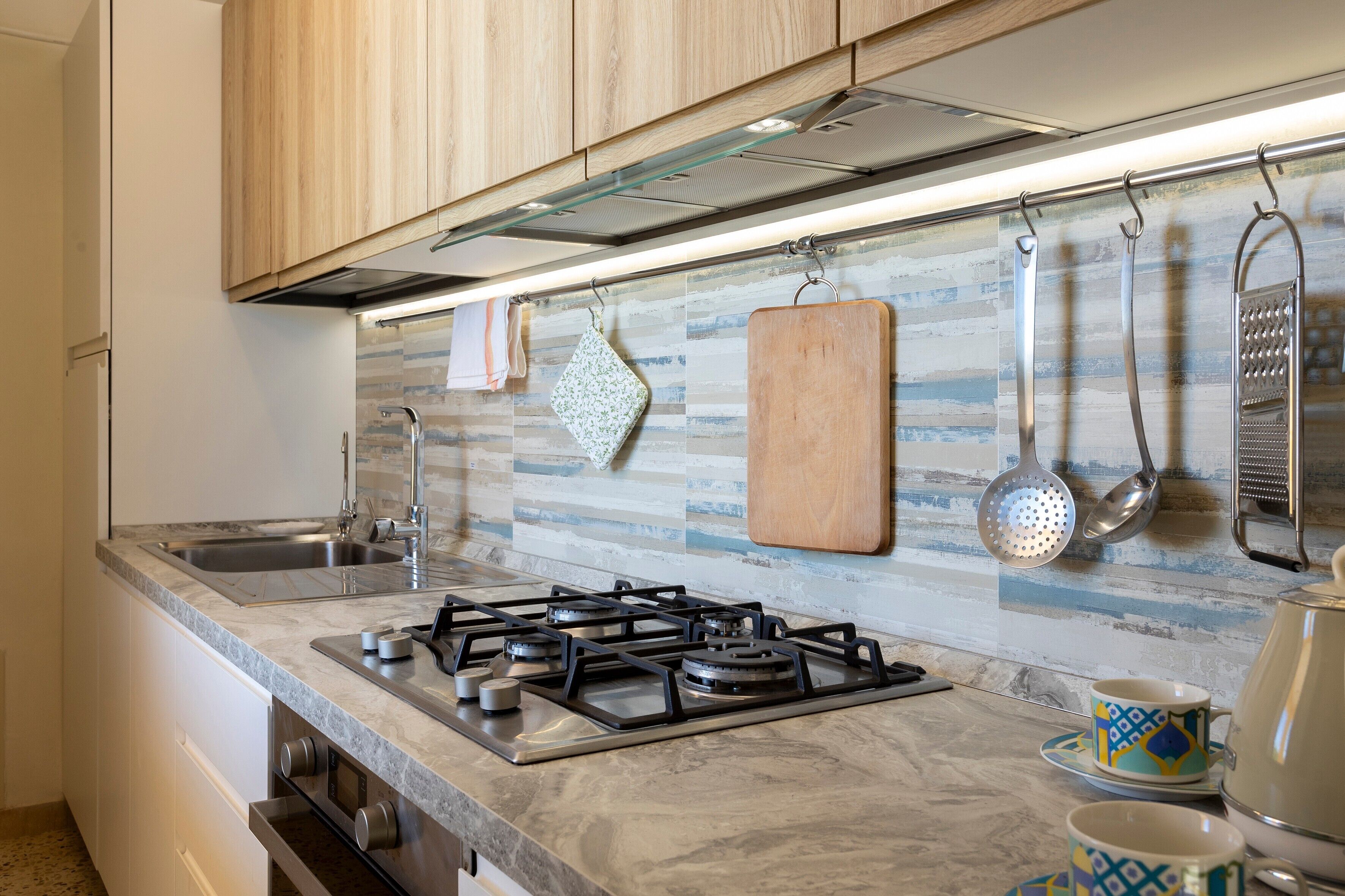 Una cocina con estufa a gas, pileta de acero inoxidable, utensilios colgantes y un salpicadero con diseño.