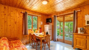 A wooden cabin with a dining table, chairs, a bed with a patterned blanket, a refrigerator, a television, and a view of greenery through the door. - Chalet 'Gite Thym' with Shared Pool, Private Terrace and Wi-Fi (Mirabel-aux-Baronnies)