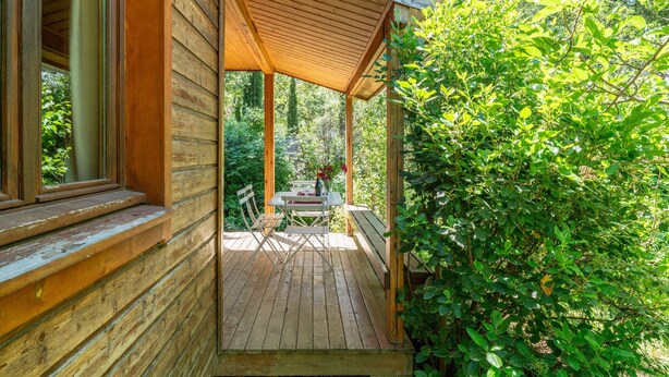 A wooden deck with a table and chairs, surrounded by greenery.