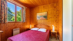 A wooden-paneled room with a bed, two bedside lamps, a painting on the wall, and a window with curtains. - Chalet 'Gite Sariette' with Shared Pool, Private Terrace and Wi-Fi (Mirabel-aux-Baronnies)