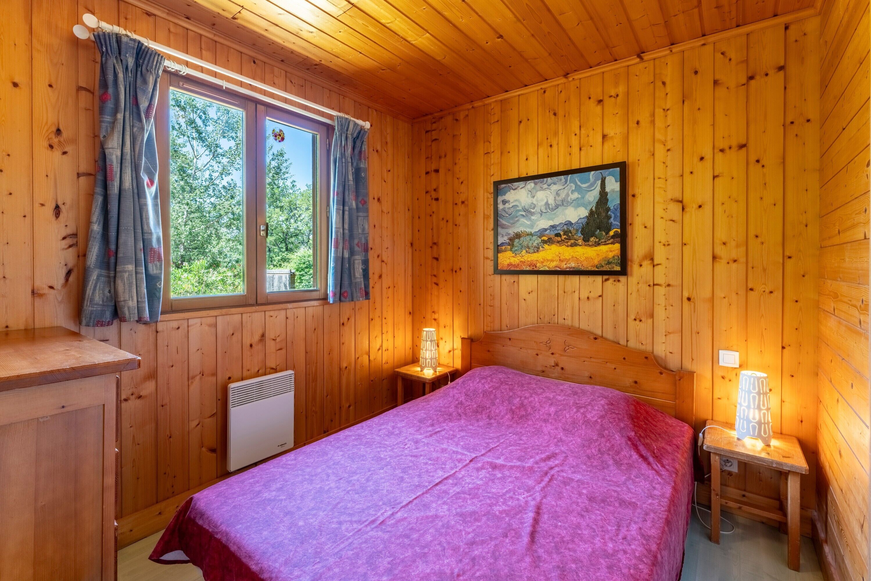Une pièce de style cabane en bois, avec un lit, une fenêtre avec des rideaux, un tableau au mur et un radiateur.