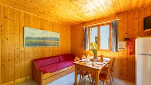 Interior de una cabaña de madera con un sofá rojo, una mesa de comedor de madera, sillas, una ventana con cortinas, un televisor montado y un refrigerador.