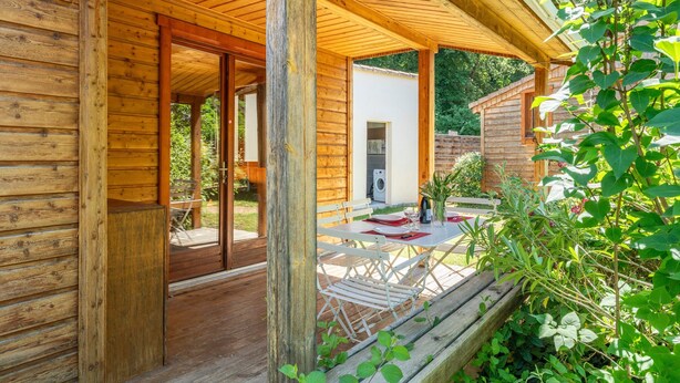 Una cabaña de madera con un porche cubierto, un comedor con mesa y sillas, y vistas del follaje circundante.
