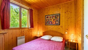 A wooden-paneled room with a bed, two bedside lamps, a radiator, and a window with curtains. - Chalet 'Gite Lavande' with Shared Pool, Private Terrace and Wi-Fi (Mirabel-aux-Baronnies)