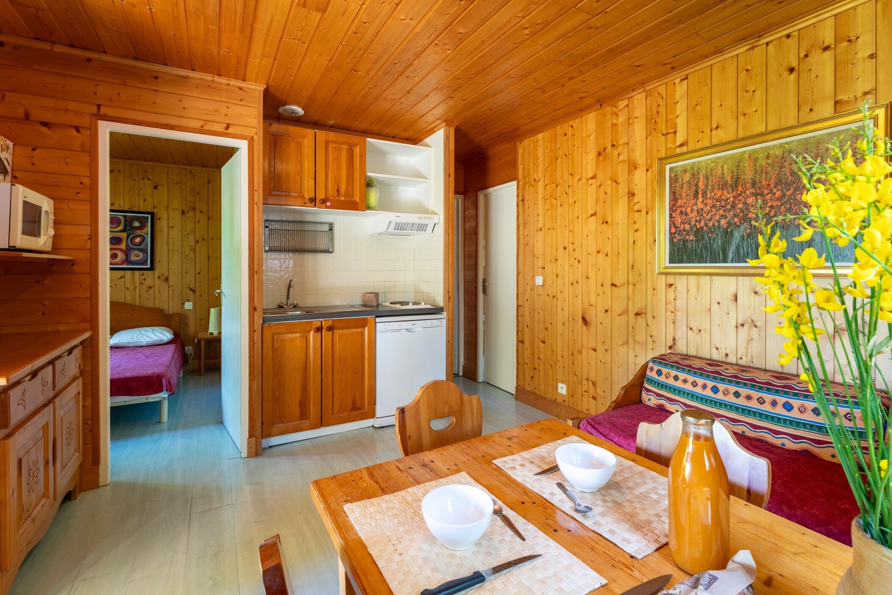 Interior de una cabaña de madera con mesa de comedor, cocina y cama con una manta colorida.