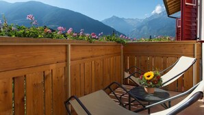 Un balcón de madera con vistas a montañas, una mesa con un arreglo floral y dos sillas.
