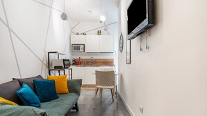 Een knus woongedeelte met een kitchenette, een bank met kussens, een eethoek en een aan de muur bevestigde televisie.