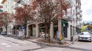 Straatbeeld met een gebouw dat een winkel op de begane grond en een restaurant met terras herbergt. Bomen met herfstbladeren, een geparkeerde auto en een voetgangersoversteekplaats.
