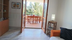 Une porte ouverte donne sur un balcon où se trouvent une table à manger et des chaises, un buffet en bois et une petite table avec une lampe.