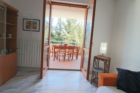 An open doorway leading to a balcony with a dining table and chairs, a wooden cabinet, and a small table with a lamp.