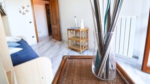 Une pièce comprenant un lit, un plateau en osier, un vase en verre avec des tiges métalliques, une petite étagère en bois et un radiateur.