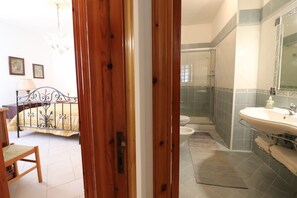 Ein Schlafzimmer mit einem Bett, einem Sessel und einer Holztür zu einem Badezimmer mit Waschbecken und Dusche.