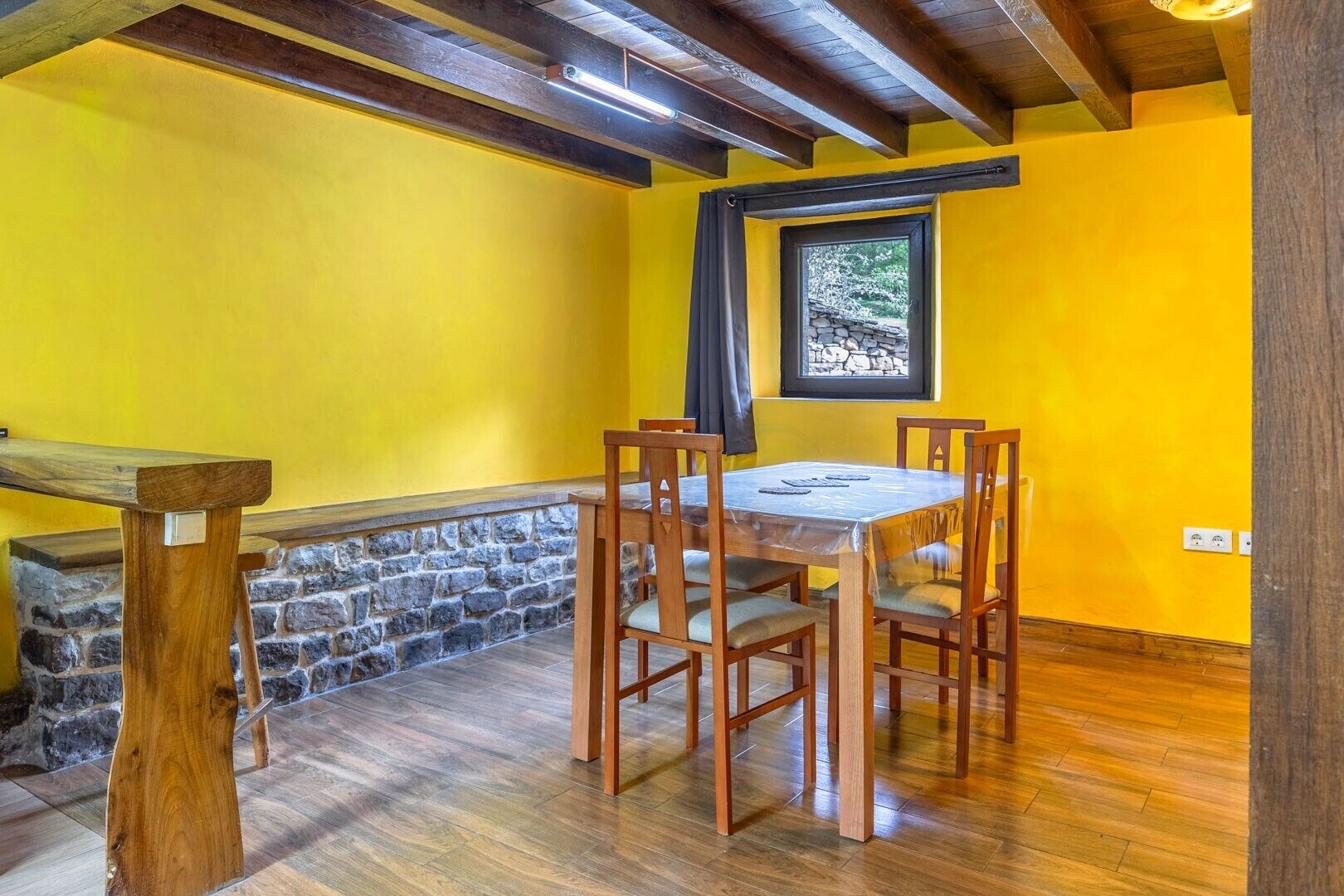 A dining area with a wooden table and chairs, a stone wall, and a window with a view of trees.