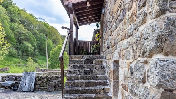 Eine Steintreppe führt zu einer Holzterrasse, begleitet von einer Steinsäule und einem grasbewachsen Hang im Hintergrund.