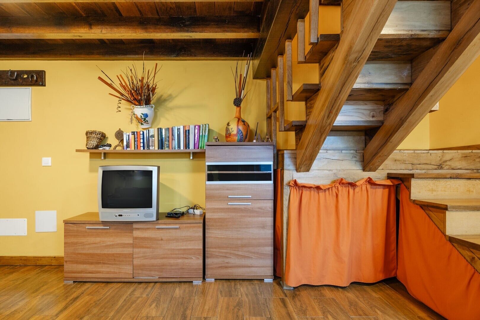 Una habitación con un mueble de televisión de madera, libros, un florero con plantas secas y una escalera.