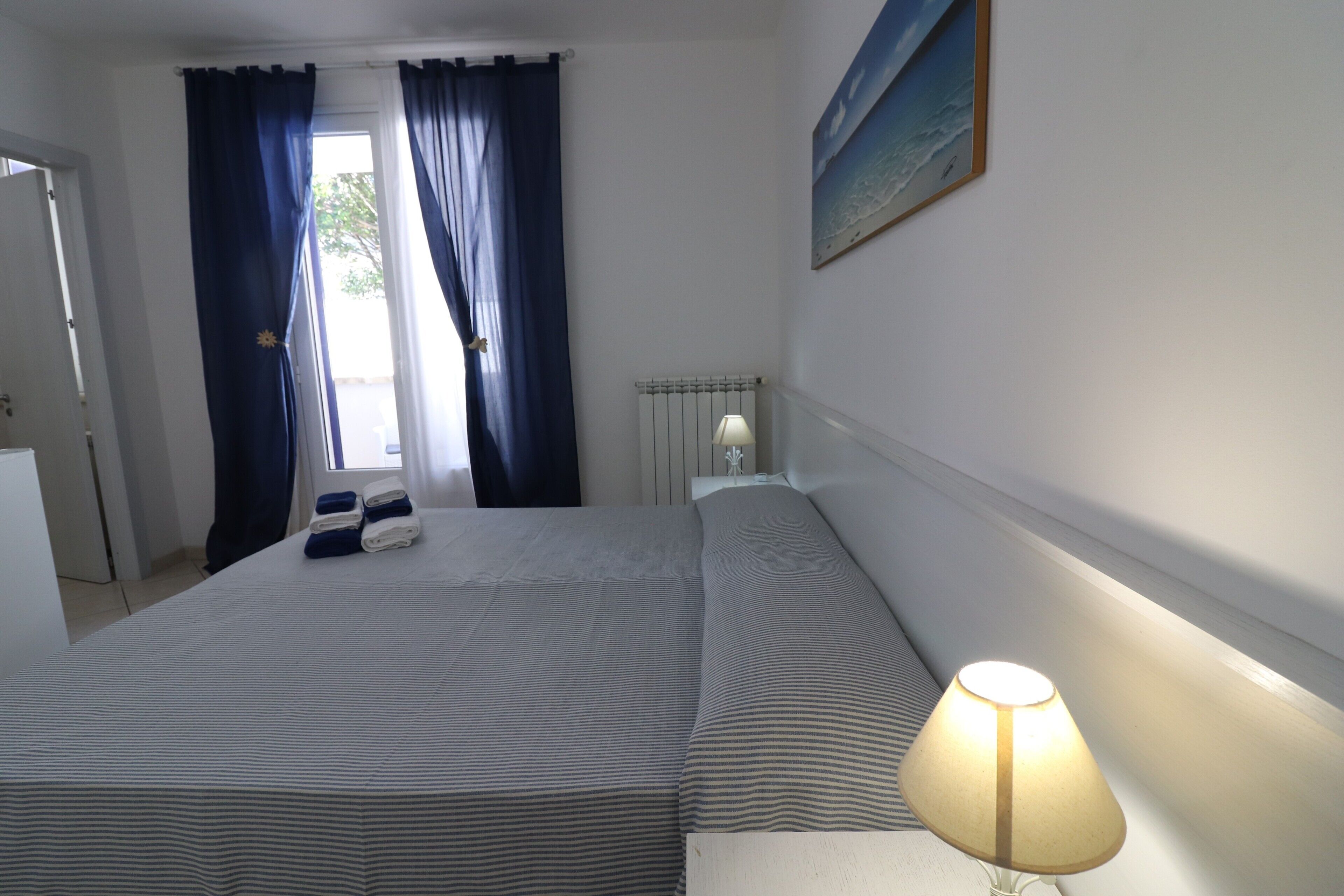 Een hotelkamer met een bed, nachtkastje, lamp, radiator en raam met gordijnen.