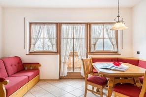 A room with a red sofa, a wooden dining table with red chairs, and a window with white curtains.