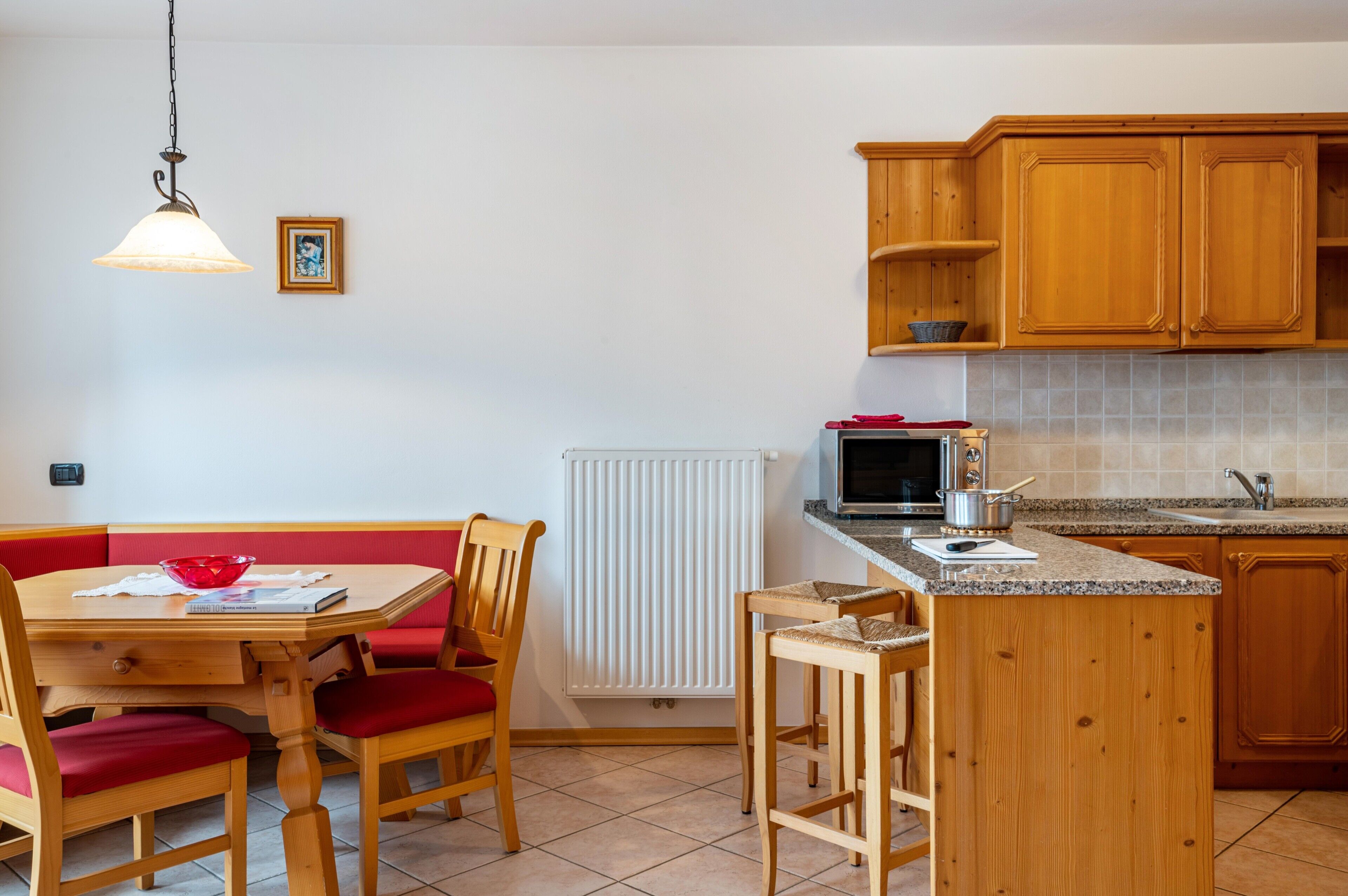 Une cuisine avec des meubles de rangement en bois, une table à manger avec des chaises rouges, un luminaire suspendu et un radiateur.
