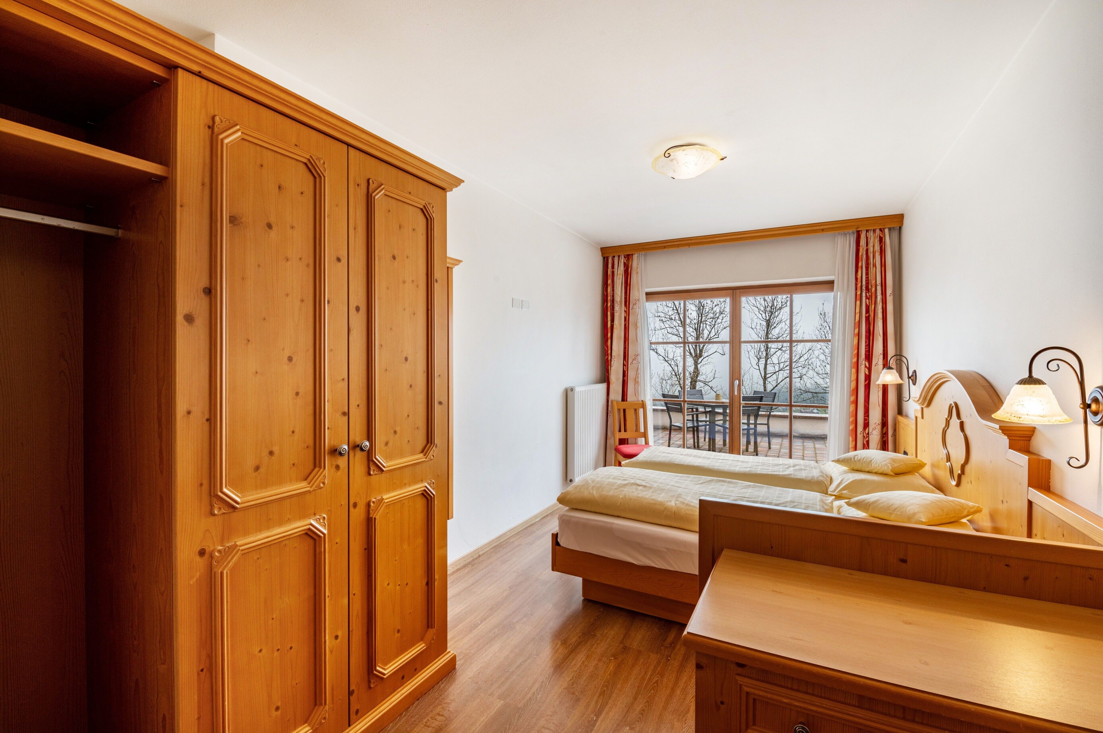 Un dormitorio con un armario de madera, una cama con almohadas, una mesita de noche, una ventana con cortinas y vistas al exterior.