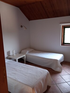 Een kleine, schone kamer met twee eenpersoonsbedden, een nachtkastje met lamp en een raam voor natuurlijk licht.