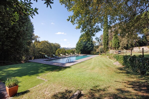 Una piscina con lettini, un prato curato e alberi circostanti.