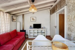 A living room with a red sofa, white armchair, glass coffee table, TV on a white cabinet, and a stone wall.