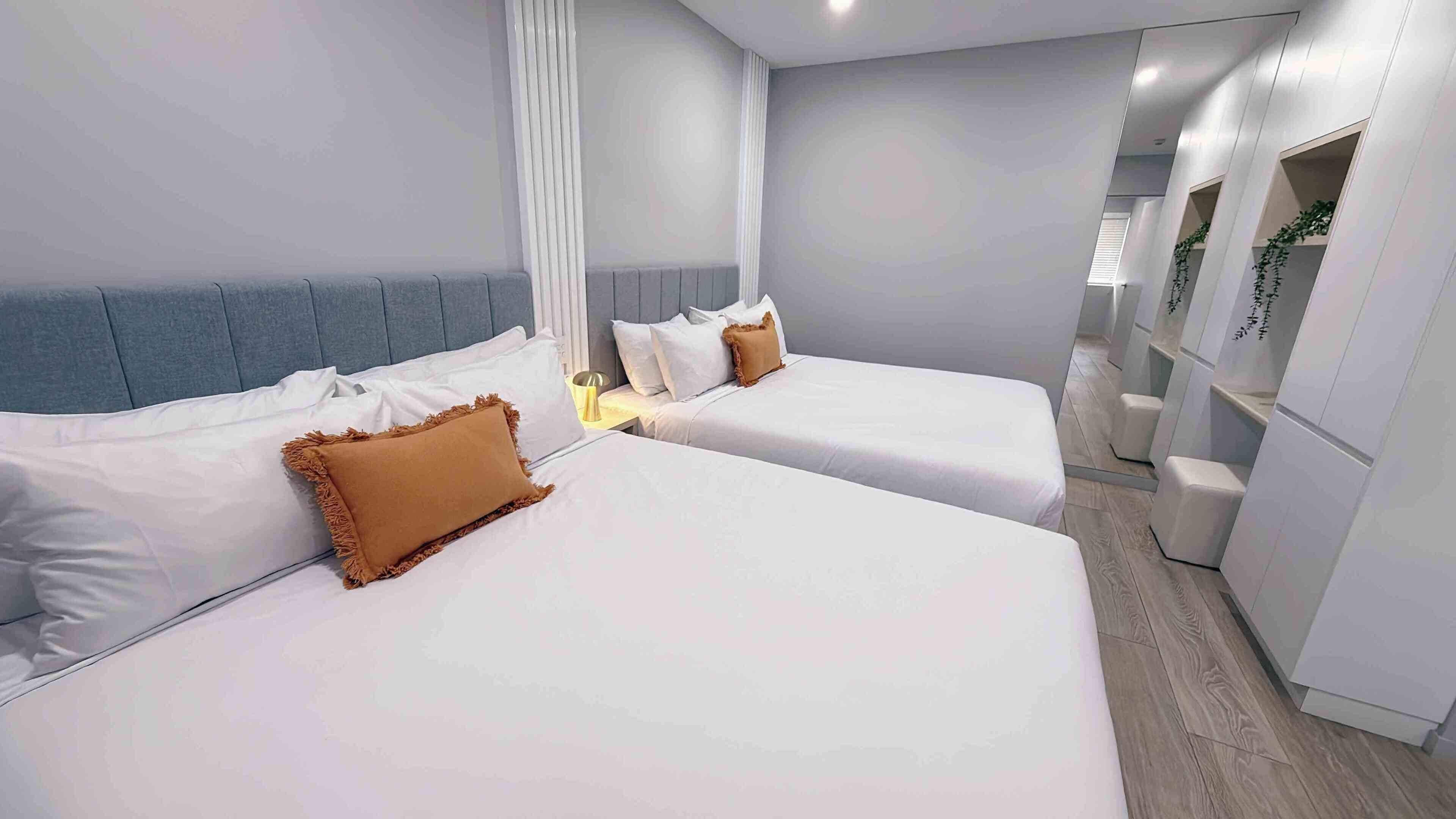 A hotel room with two beds, a grey headboard, white bedding, and a small bedside table with a lamp.
