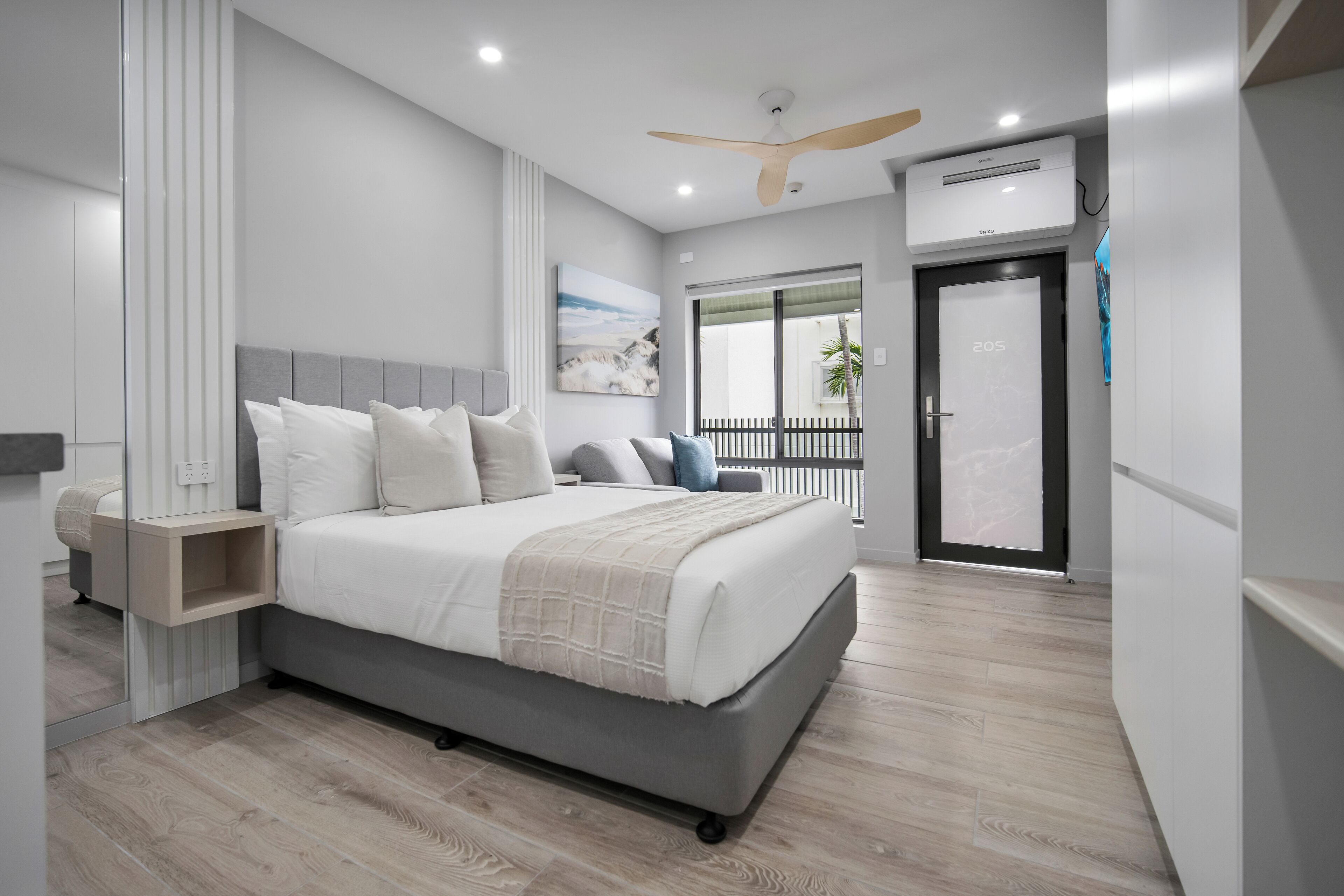 Quarto moderno com uma cama grande, mesas de cabeceira, ventilador de teto, ar condicionado e uma porta de vidro deslizante para uma varanda.